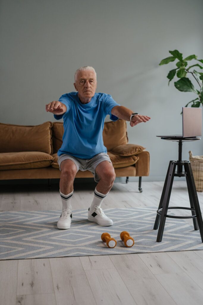 An older man doing a bodyweight squat at home following a workout on a laptop